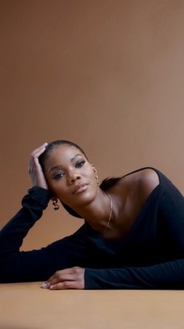 A Black woman in an off-shoulder black top reclines gracefully, wearing gold jewelry against a warm brown background.