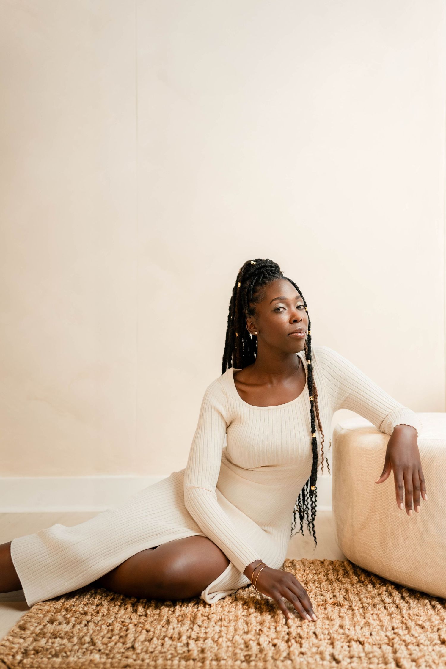 A young Black woman with braided hair sits gracefully on a woven rug in a cream ribbed knit dress, leaning on a beige chair.
