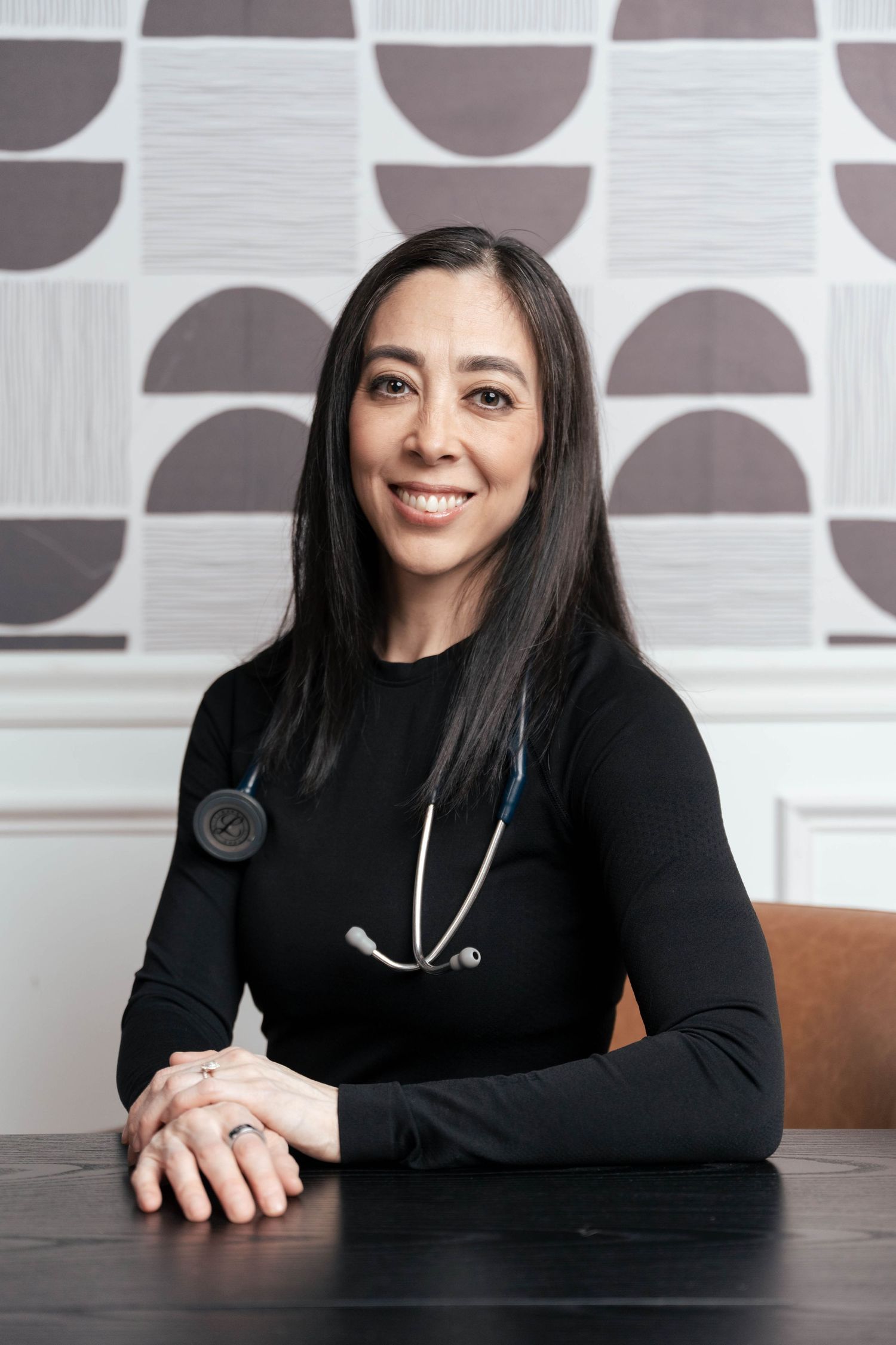 Smiling female doctor in black top with stethoscope seated at desk with geometric wallpaper background.