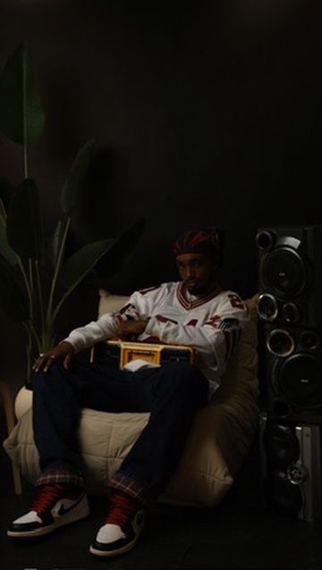 Man in white jersey, jeans, and Air Jordan 1s sits in chair holding boombox beside speaker and plant.