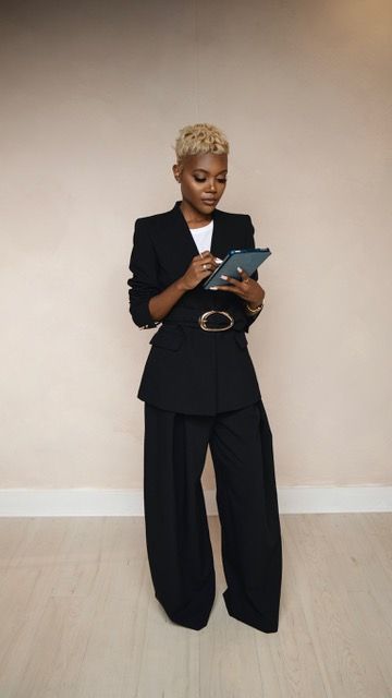 A confident woman in a black blazer and wide-leg pants holds a tablet, wearing a gold belt and blonde cropped hair.
