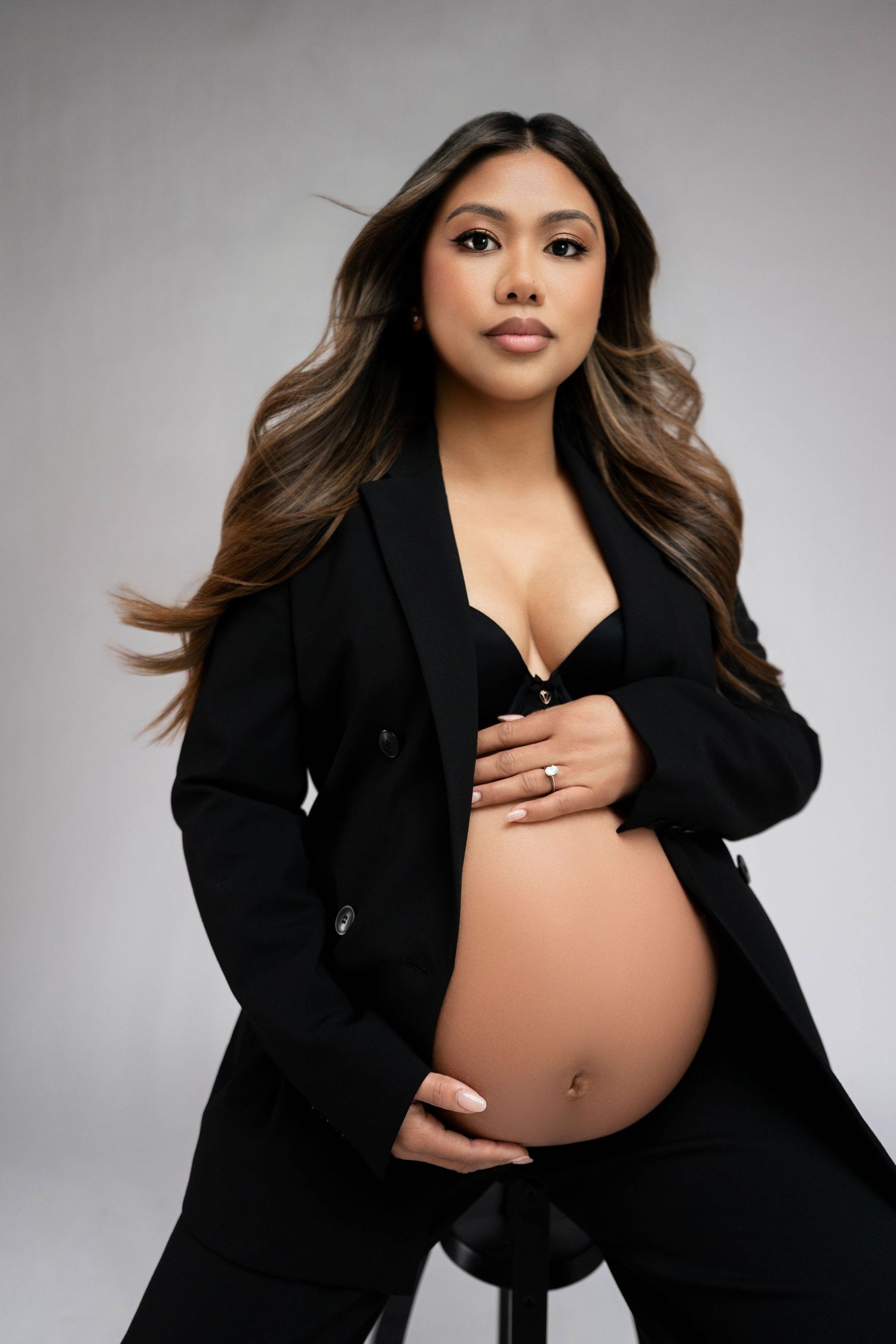 Pregnant woman in black blazer and bra cradling baby bump, with long wavy hair, posing in studio.