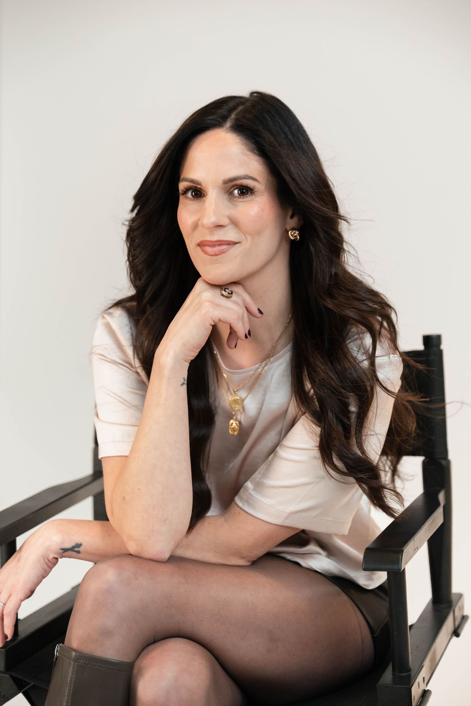 Woman with dark wavy hair sits cross-legged on a director's chair, wearing a white top and gold jewelry.