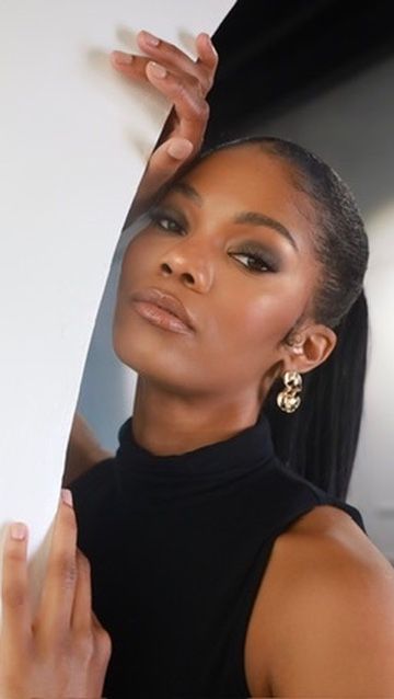 A Black woman with sleek hair poses elegantly in a black turtleneck, wearing gold hoop earrings beside a white panel.