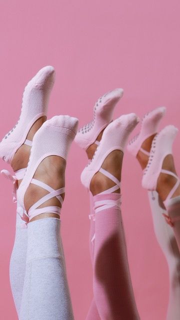 Multiple legs raised wearing pink ballet-style grip socks with criss-cross ankle straps on pink background.