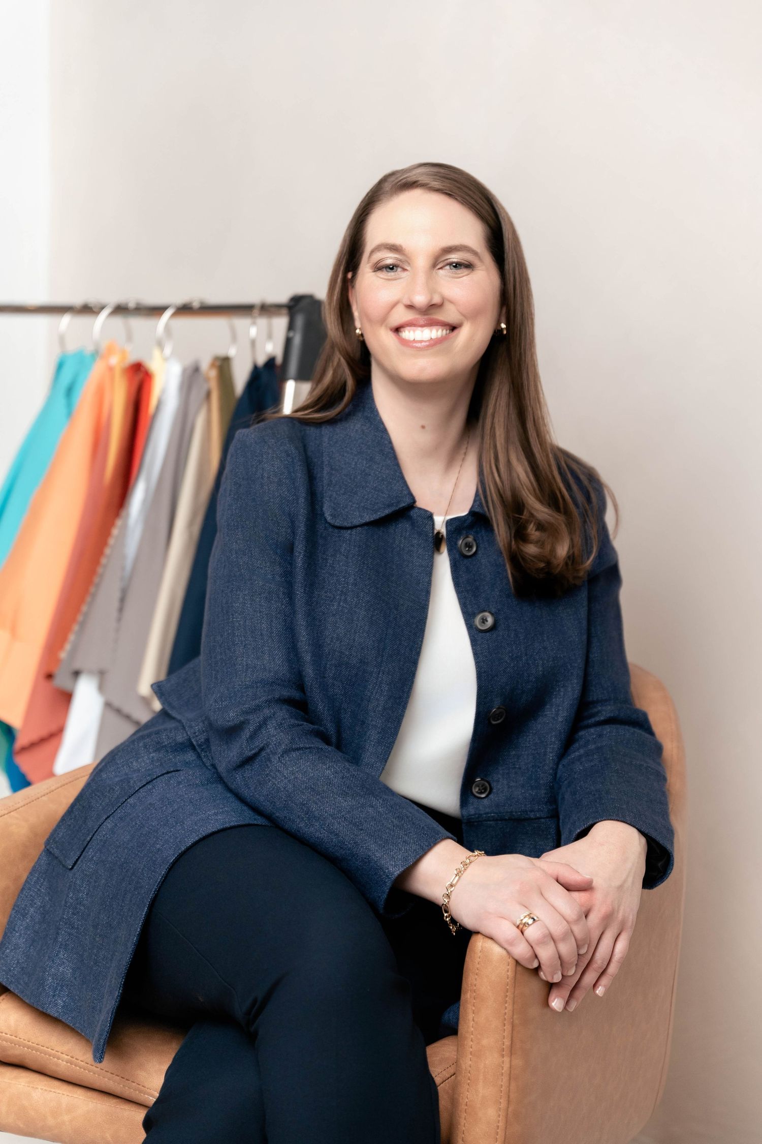 Smiling woman in navy blazer sits in leather chair before clothing rack in professional portrait photo.