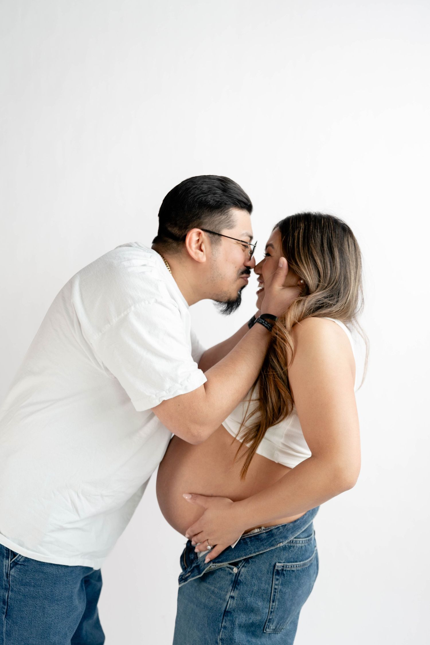 Couple in white tops and jeans sharing a tender moment, woman showing pregnant belly, maternity photo session.