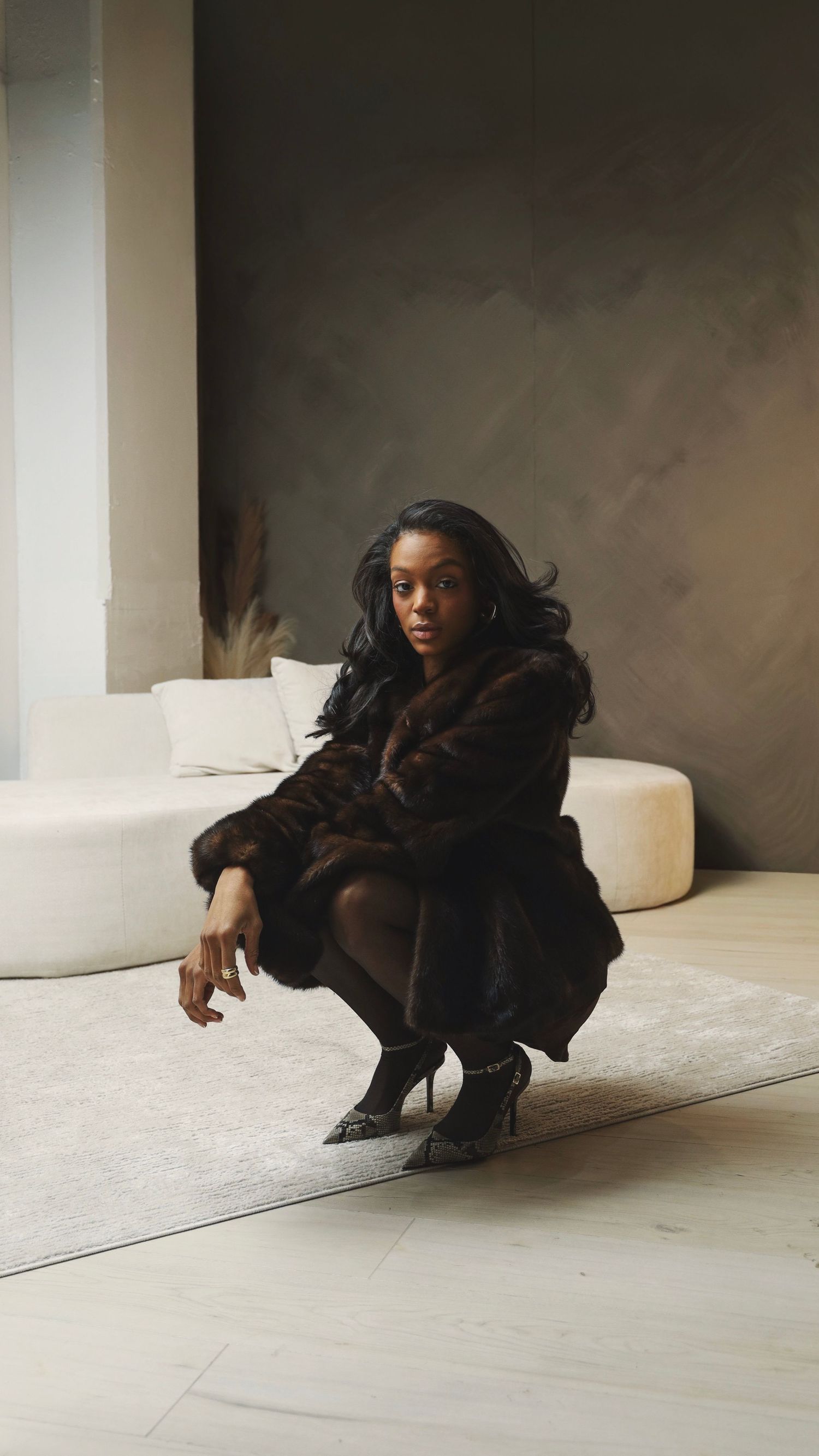 Woman in dark fur coat and heels crouching on white rug in modern minimalist interior space.