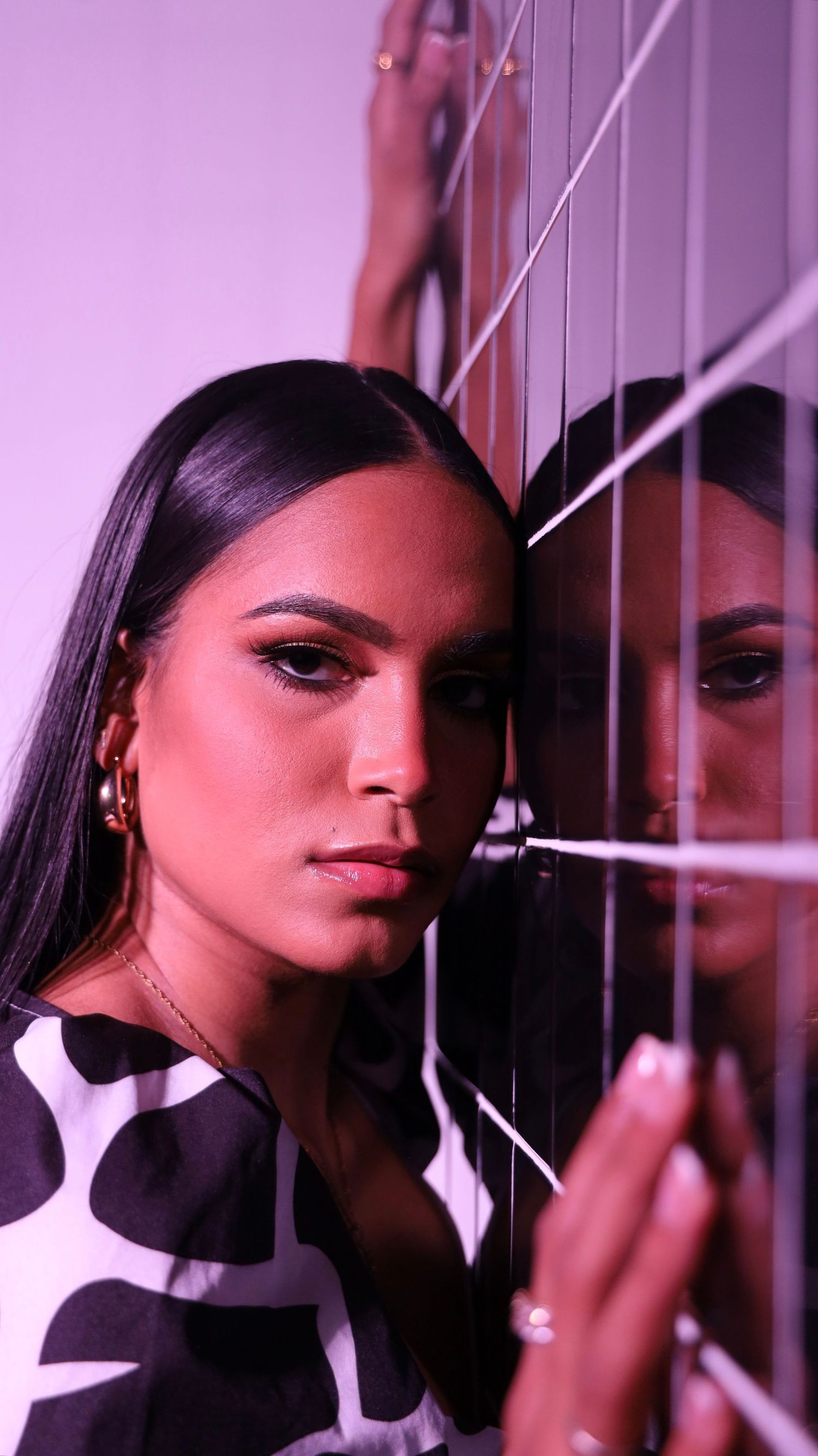 Woman with sleek hair and gold earrings leans against a mirrored surface in purple-toned light.