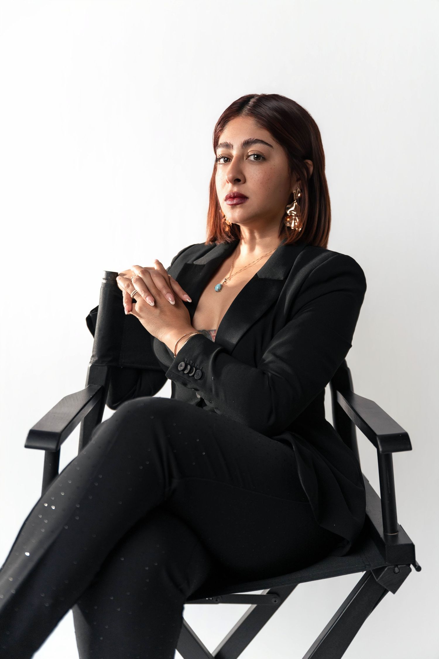 Woman in black blazer and pants sits confidently in director's chair against white background, wearing gold earrings.