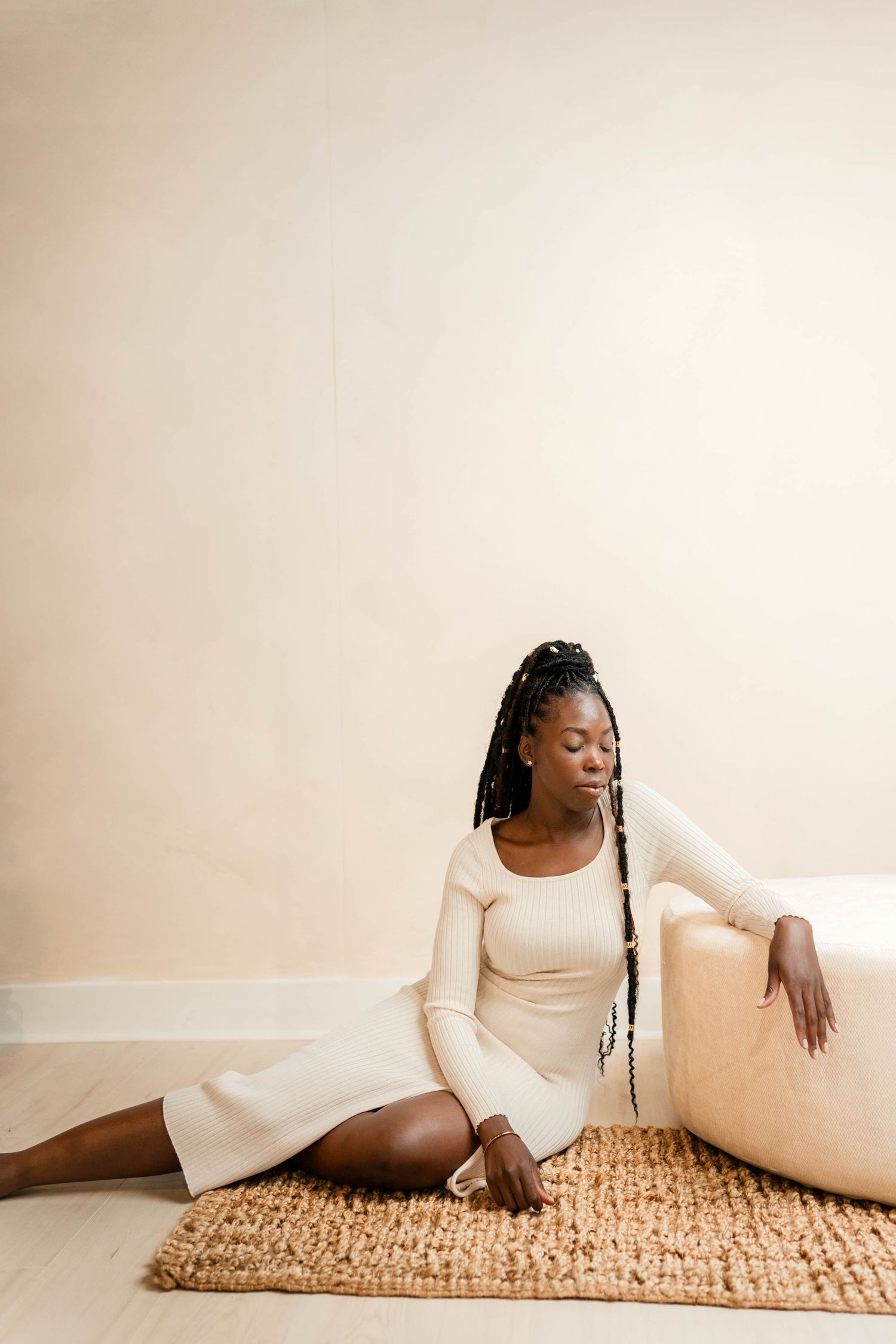 A young Black woman with box braids sits gracefully on a woven rug in a cream ribbed dress, leaning against a beige sofa.