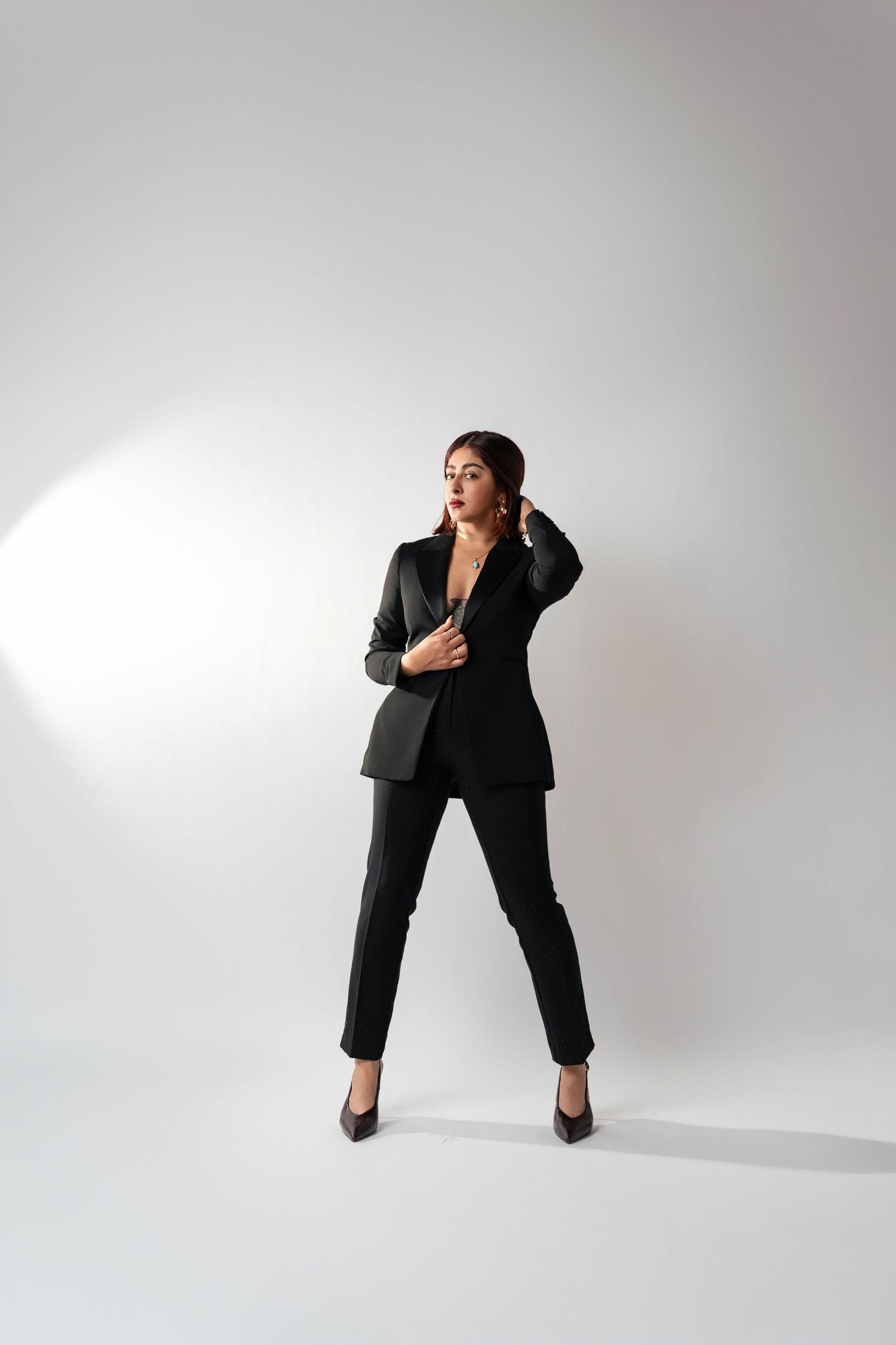 Woman in black blazer and trousers posing confidently on white background wearing black heels.