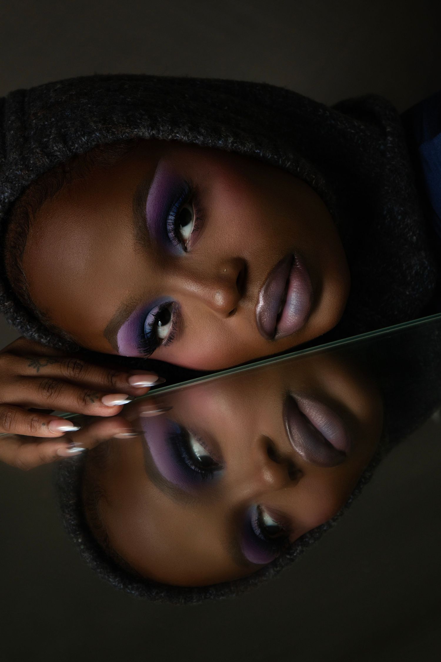 A Black woman with purple smoky eye makeup rests her face against a mirror, reflecting her beauty in dramatic studio lighting.
