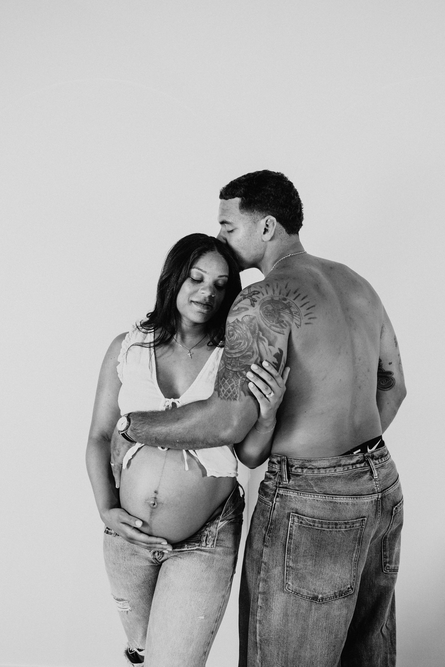 Black and white maternity photo of a pregnant woman and shirtless tattooed man embracing in jeans.