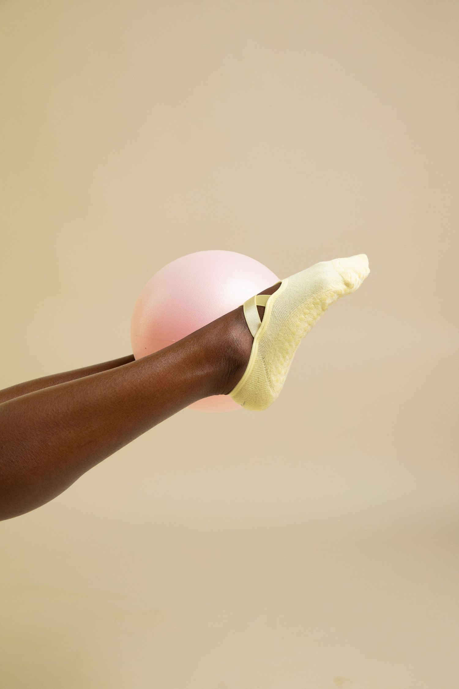 A dark-skinned foot wearing a yellow sandal balancing a pink ball against a beige background.
