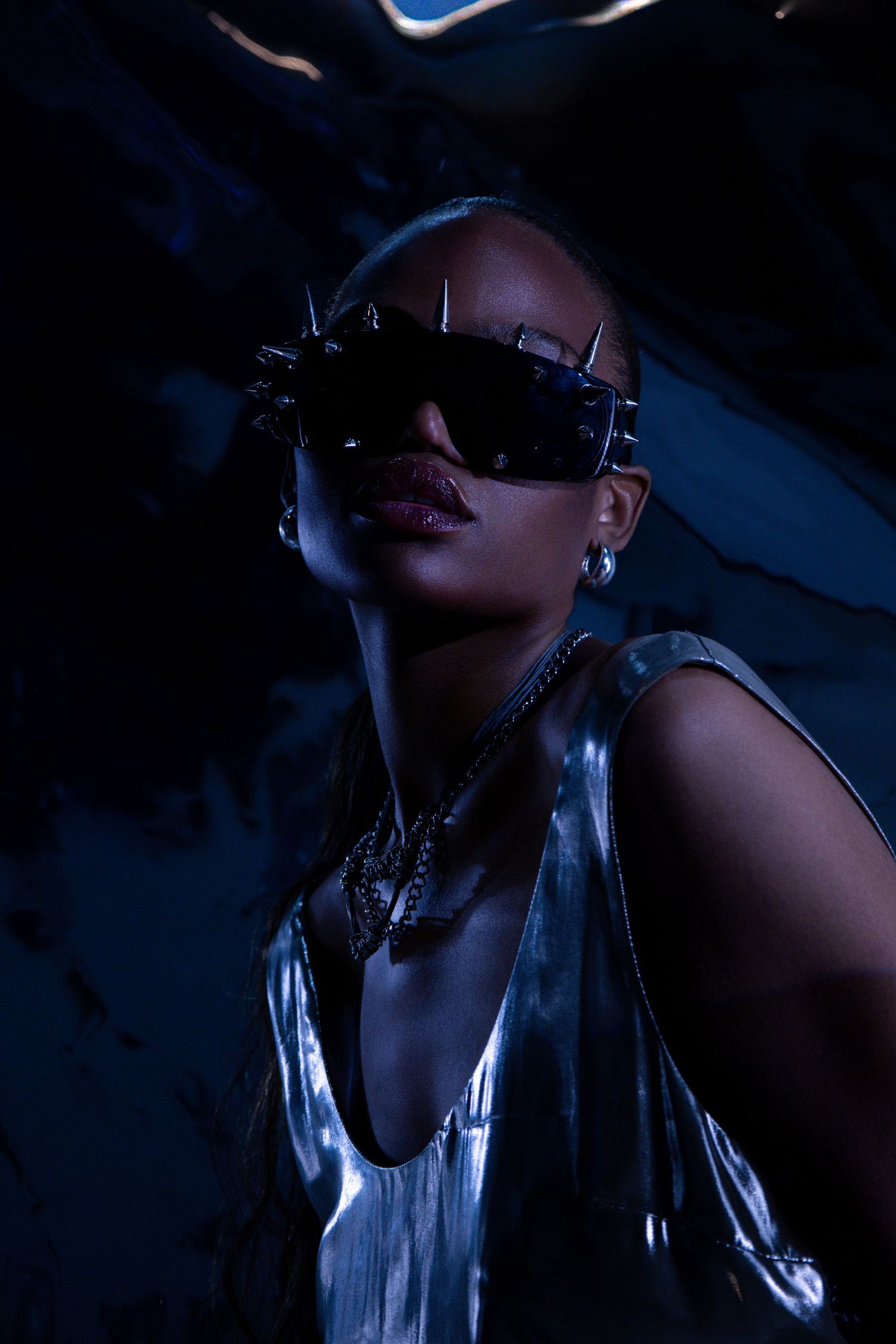 Woman in silver metallic top and spiked sunglasses poses dramatically under blue lighting.