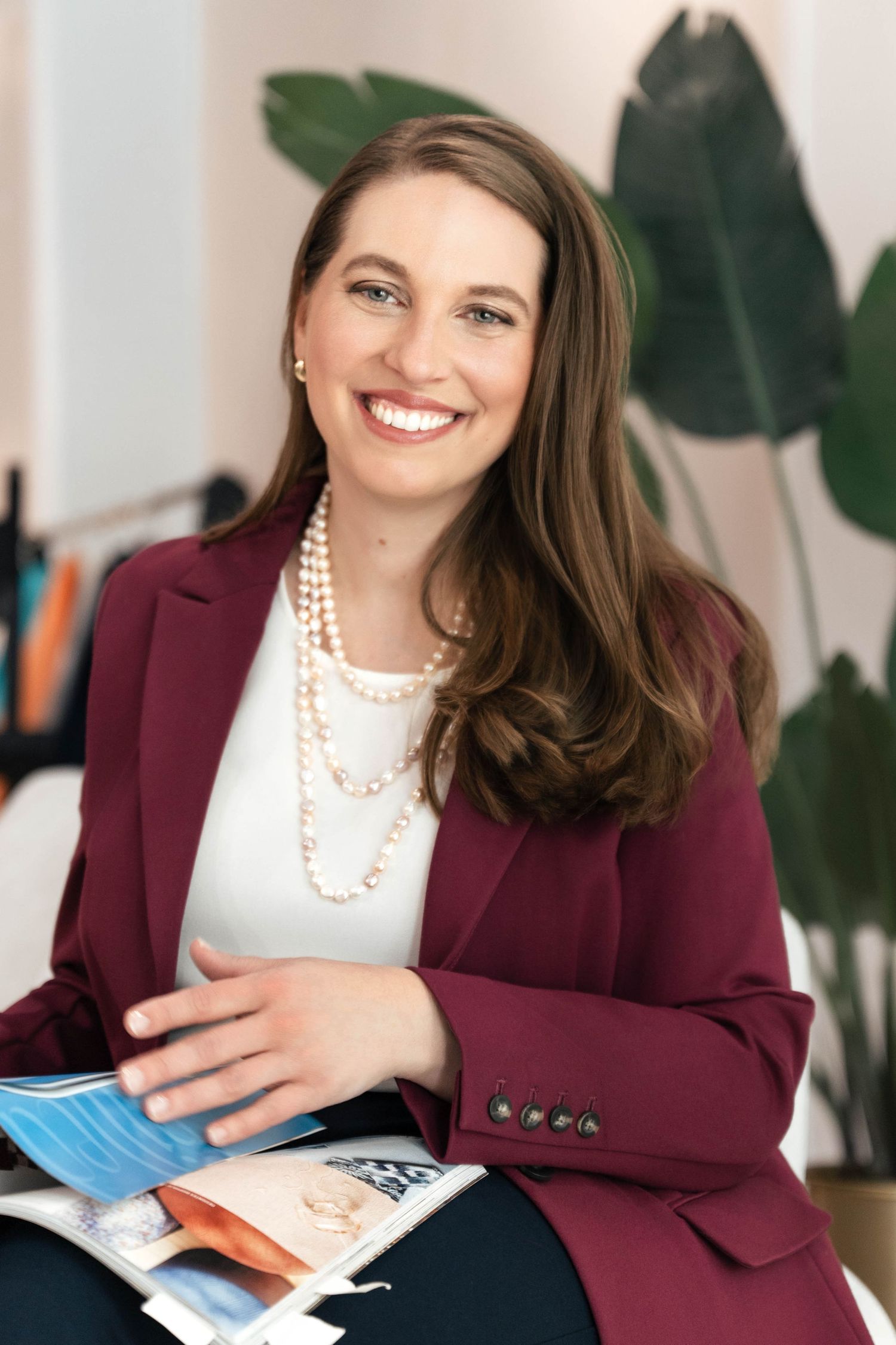Professional woman in burgundy blazer and pearl necklace smiling while holding a magazine in a bright office.
