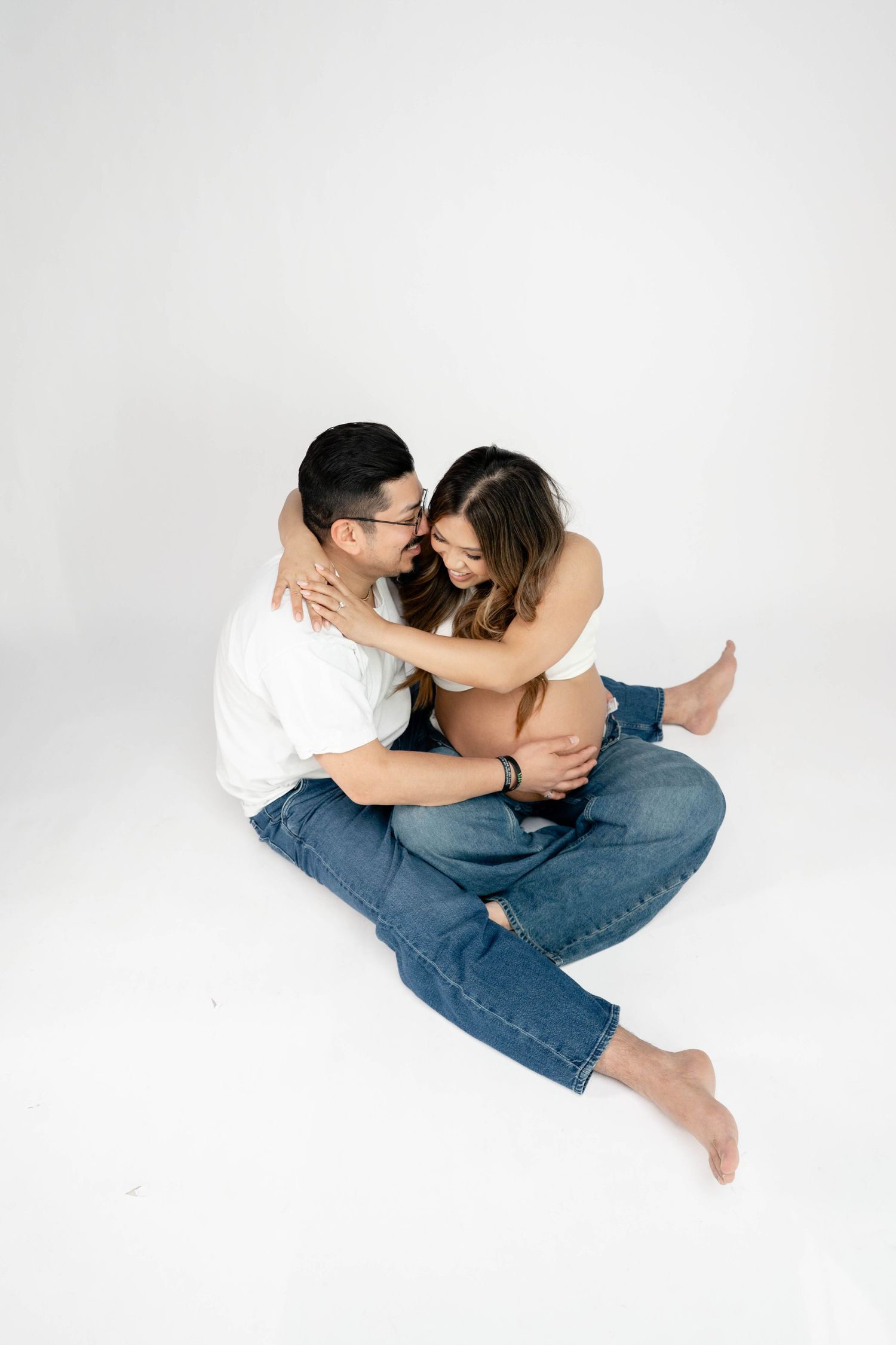 Couple sitting on white floor embracing, wearing jeans and casual tops, sharing a tender romantic moment.