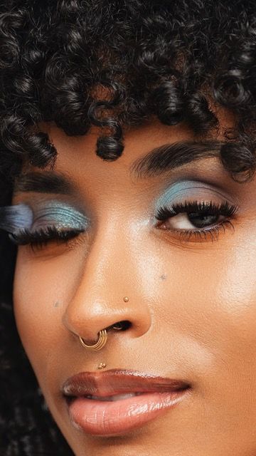 Close-up of woman with blue eyeshadow, gold nose ring, and curly hair displaying a stunning makeup look.