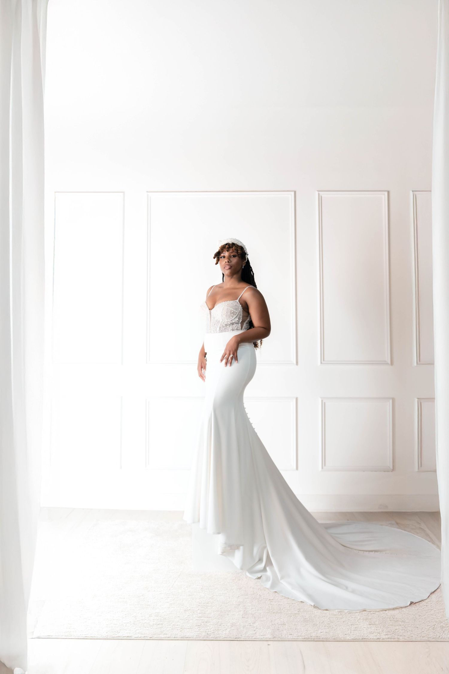 Elegant Black bride in a white mermaid wedding gown with a long train and floral crown in a bright room.
