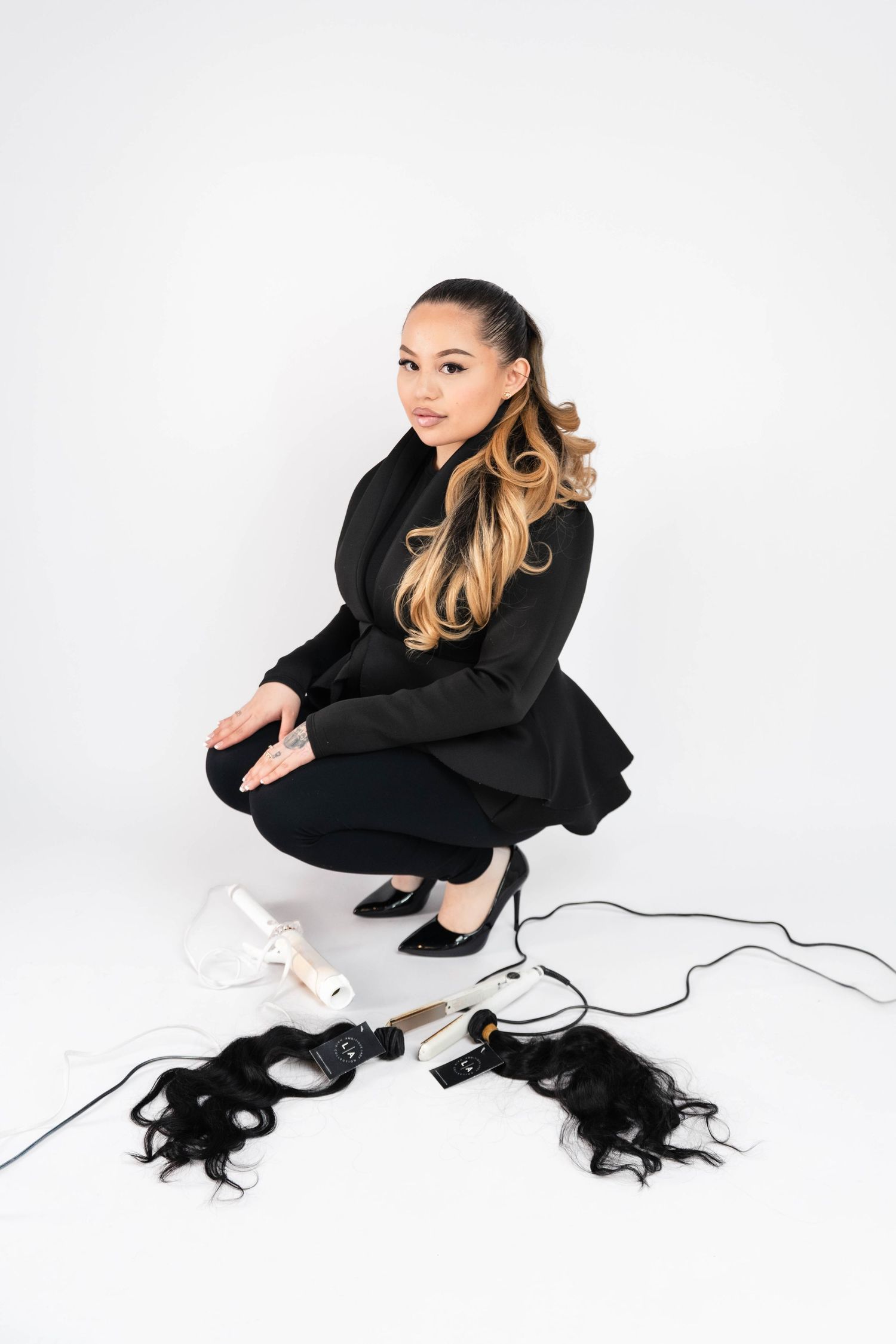Woman in black outfit crouching beside hair tools, curling iron, and extensions on white background.