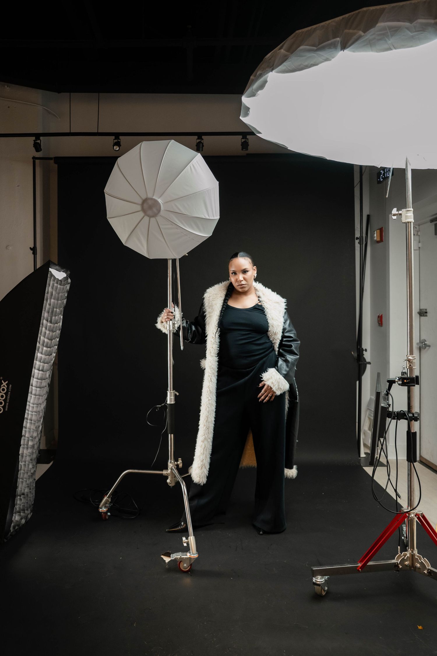 Woman in black outfit and white fur coat poses in photography studio with umbrella lights and black backdrop.