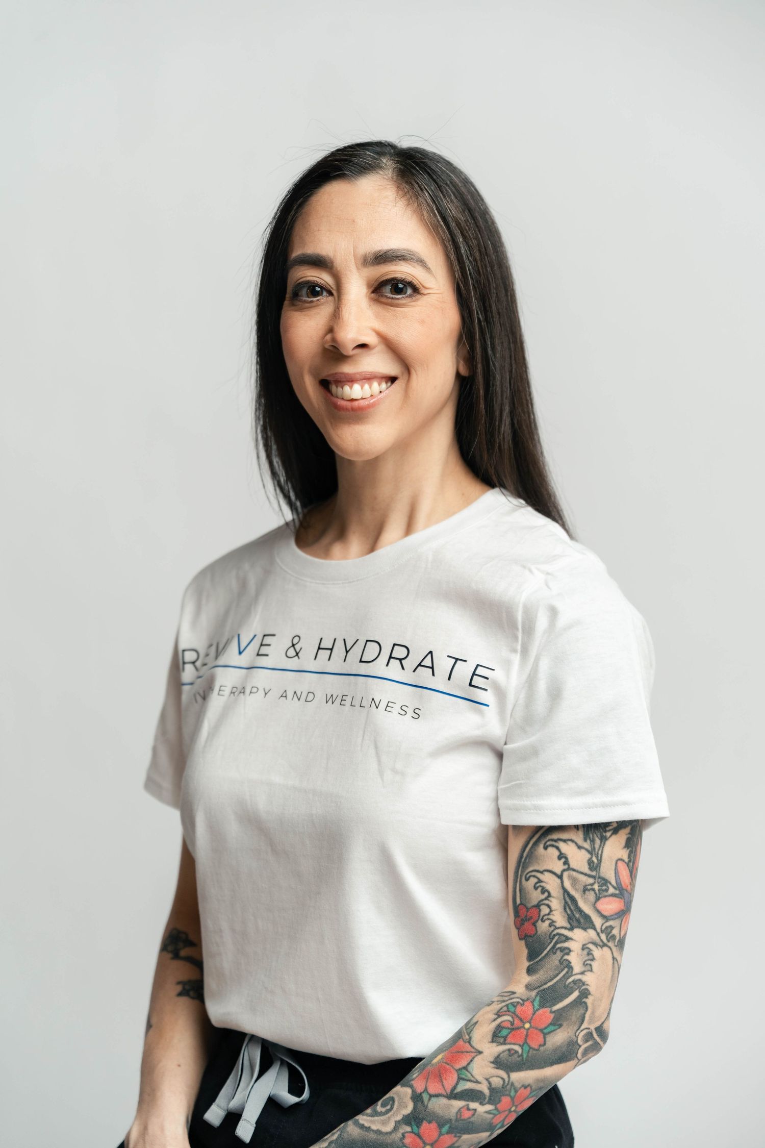 Smiling woman with long dark hair and sleeve tattoo wearing a white Revive & Hydrate wellness t-shirt.