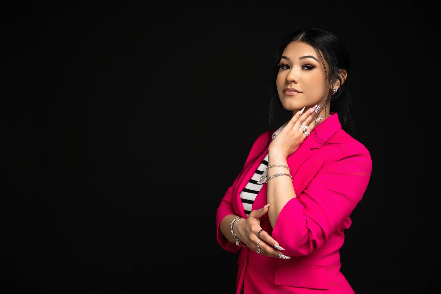 Woman in pink blazer posing confidently against black background, touching chin with manicured nails.