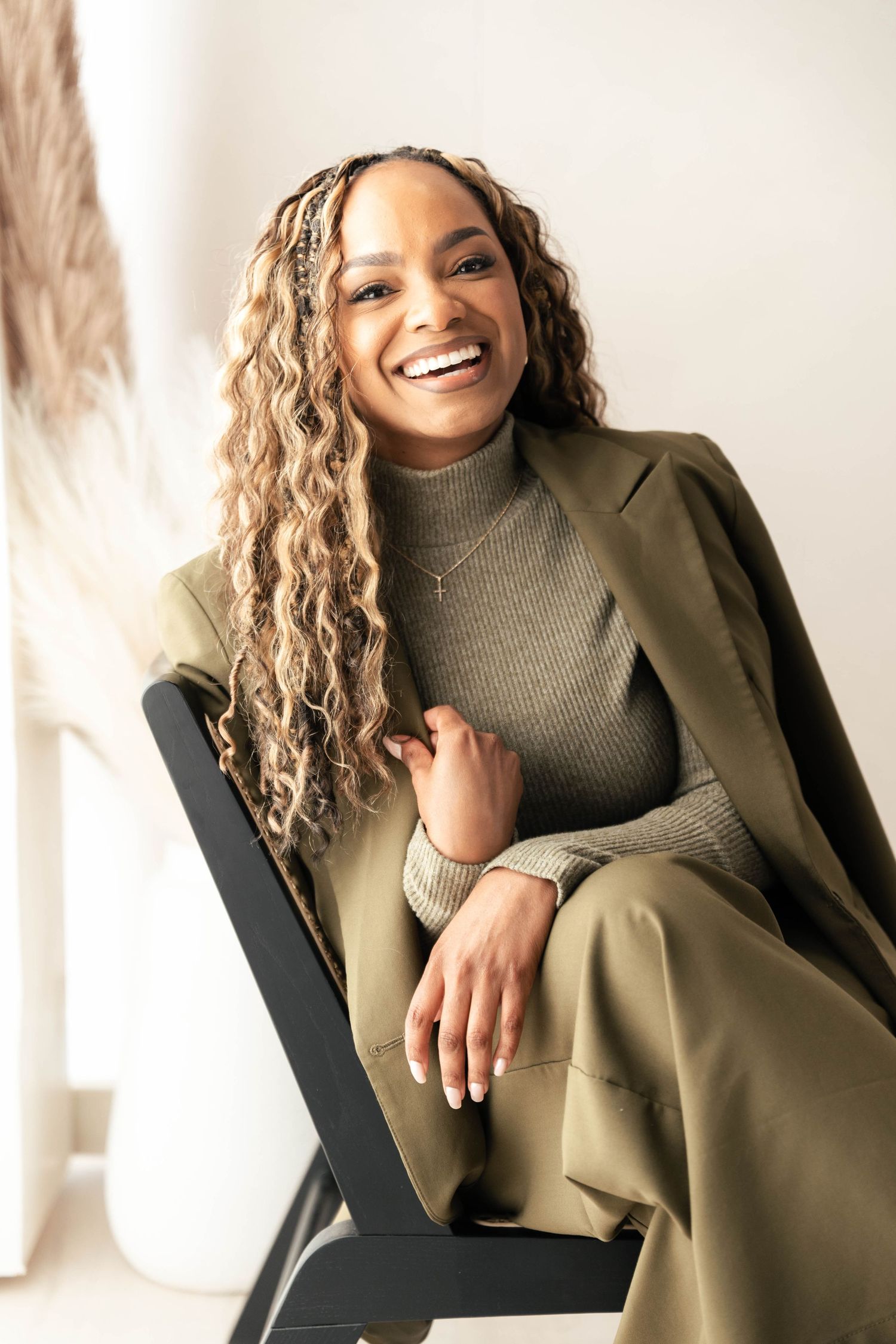 Smiling woman in olive green suit with curly blonde hair seated in black chair against white background.