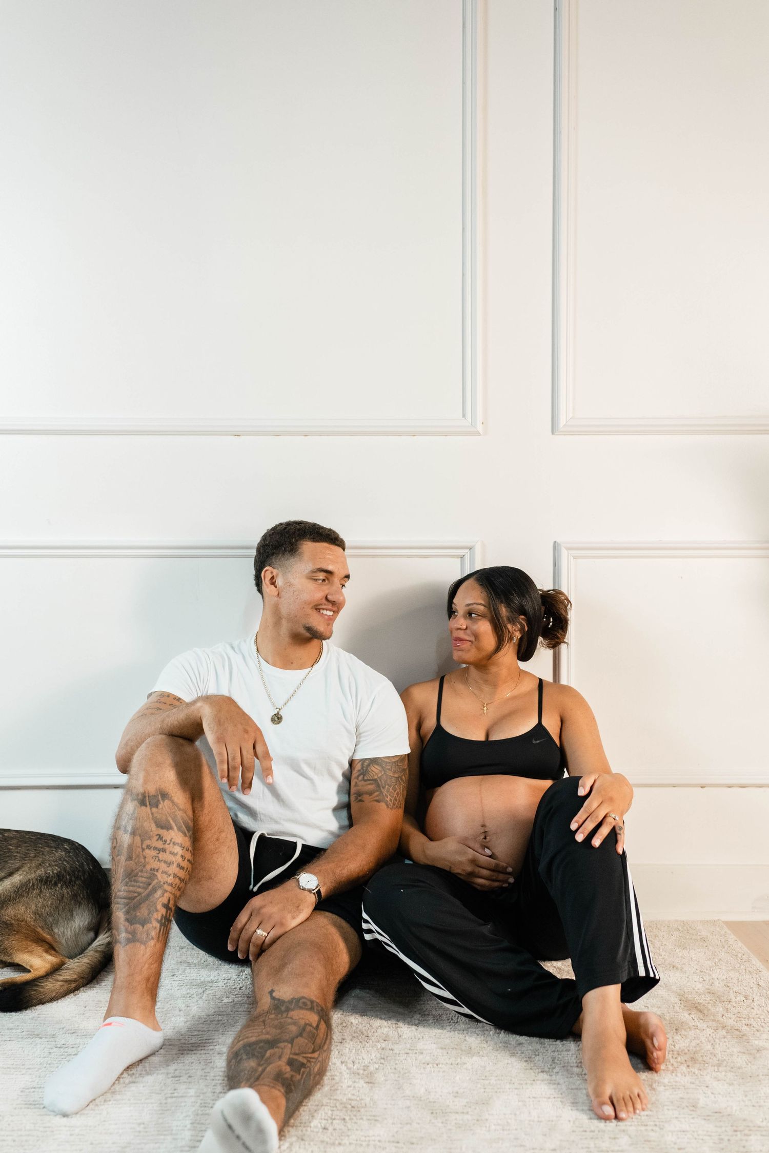 A tattooed man and pregnant woman sit together on the floor, smiling at each other with a dog resting nearby.