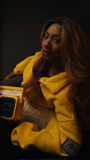 Woman with wavy auburn hair poses confidently in a bold yellow jacket holding a vintage boombox.