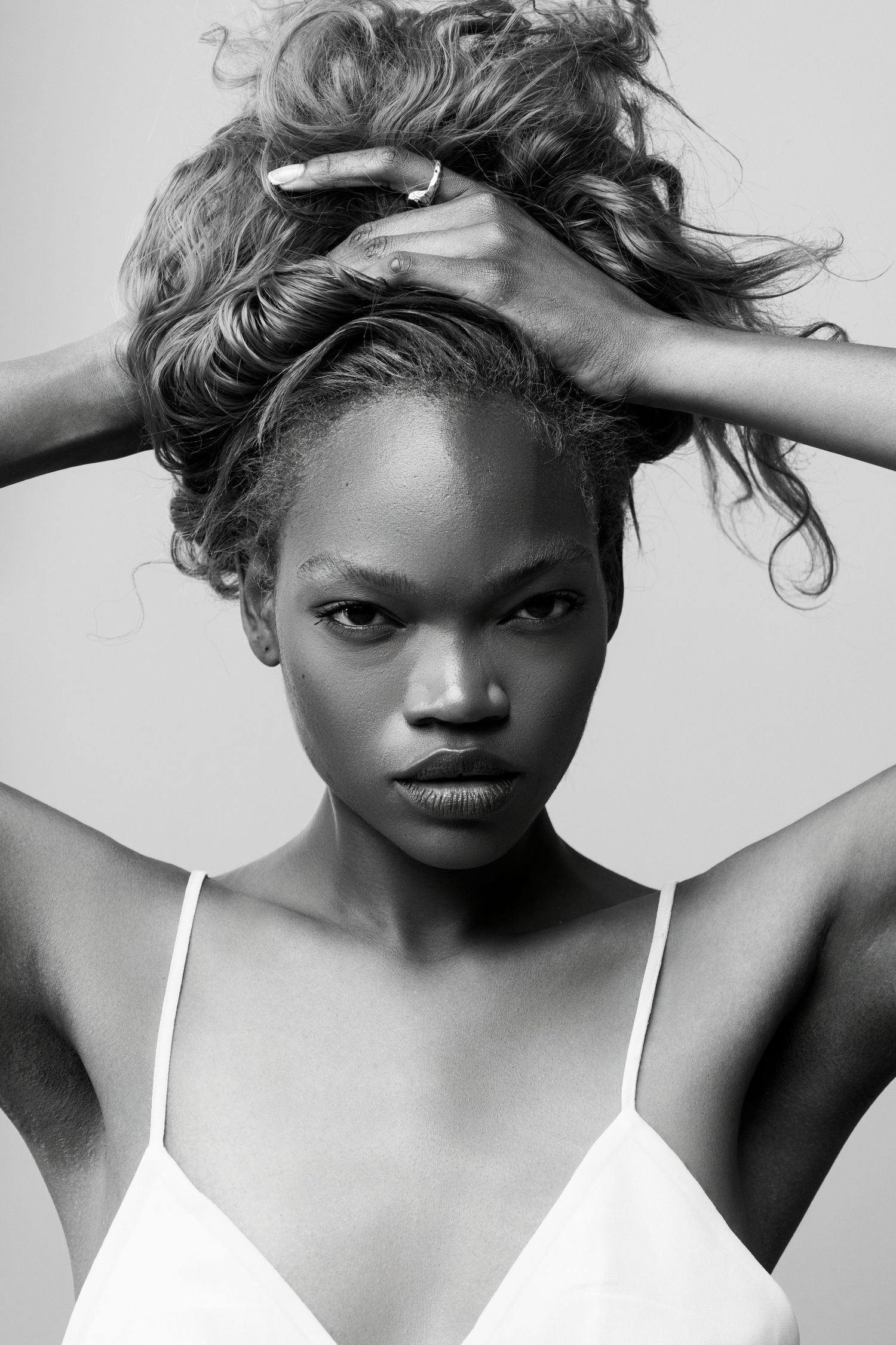 Black and white portrait of a young woman holding her curly hair up, wearing a white top, against a light background.