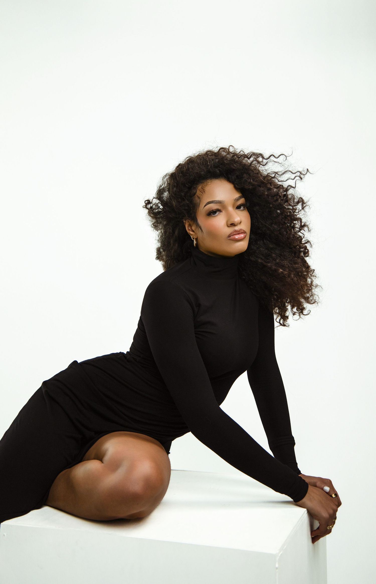 A young Black woman in a black turtleneck dress poses gracefully on a white cube with curly hair flowing.