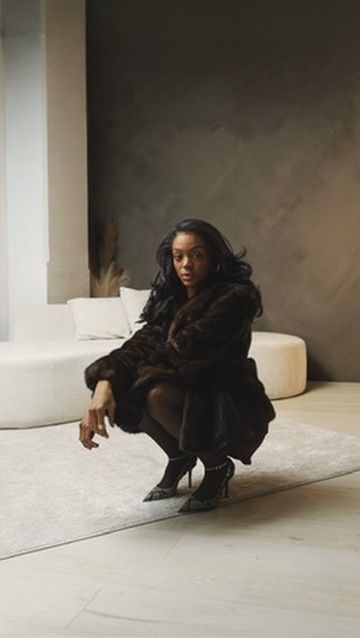 Woman in dark brown fur coat crouching on white tile floor in a modern minimalist interior.