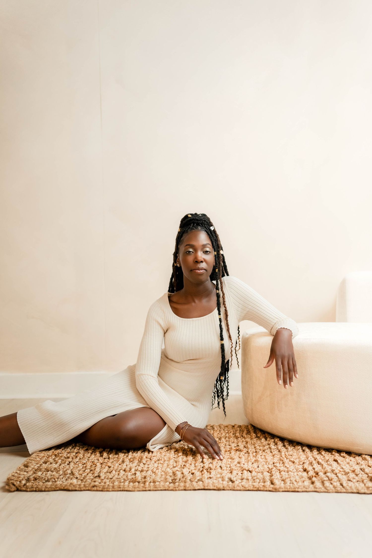 A Black woman with long braids sits gracefully on a woven rug in a cream ribbed dress, leaning against a beige sofa.