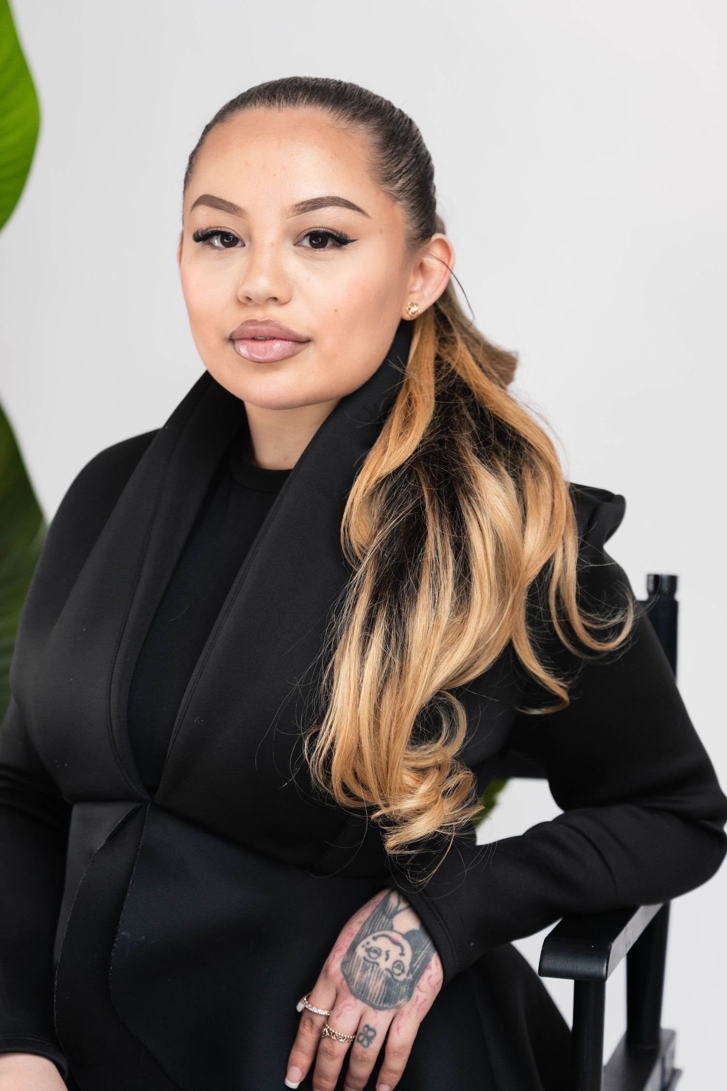 Young woman in black blazer with ombre hair and hand tattoo poses confidently against white background.
