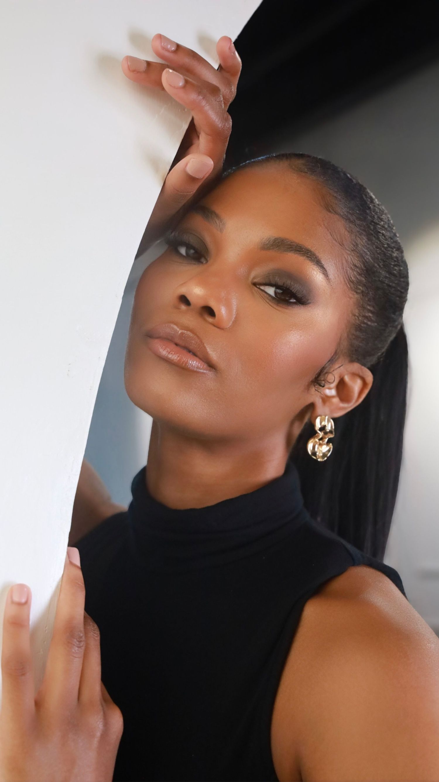 A Black woman in a black turtleneck poses with a mirror, wearing gold earrings and smoky eye makeup.