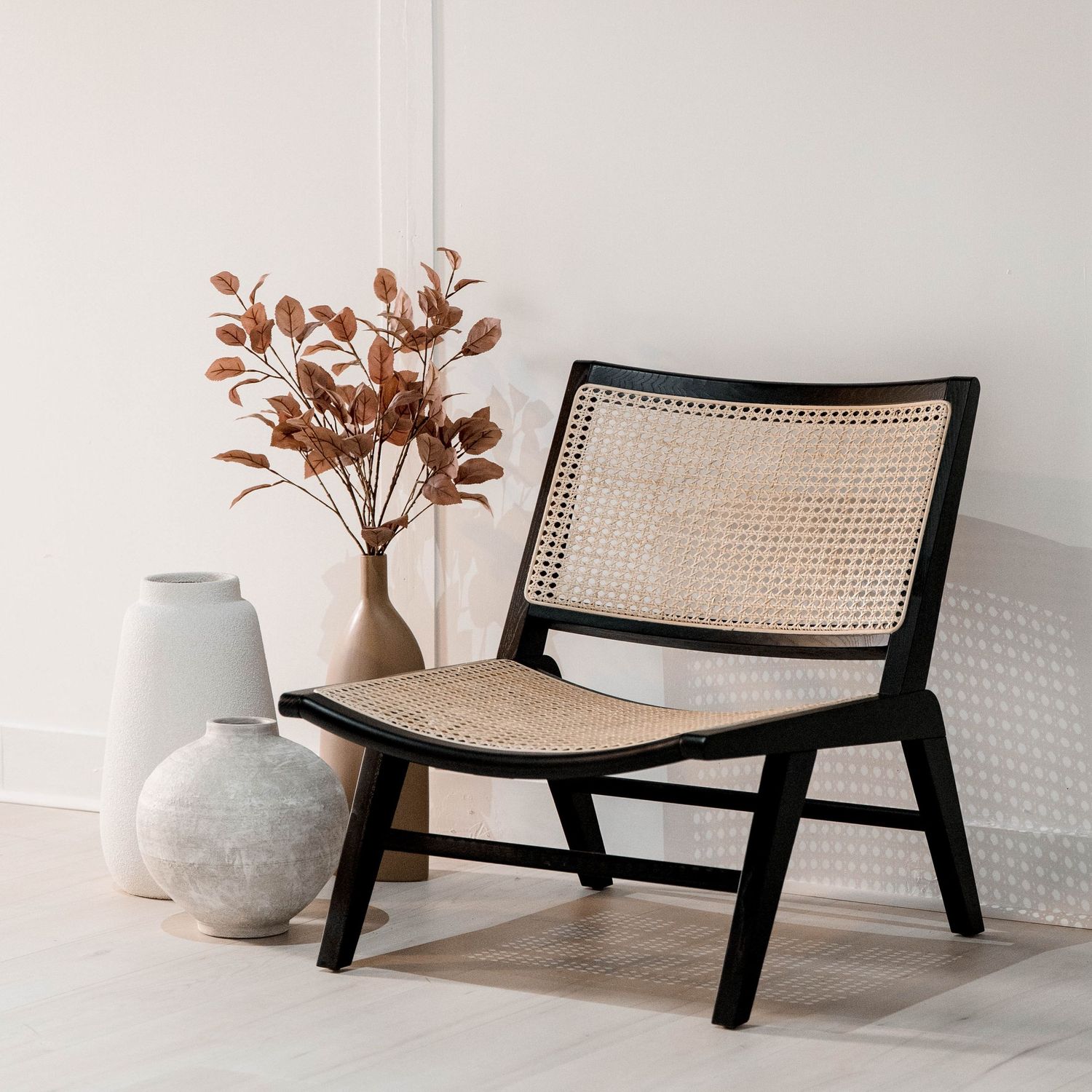 Modern black-framed rattan lounge chair with dried flowers in a ceramic vase on light wooden flooring.