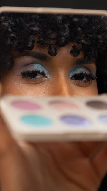 Woman with blue eyeshadow holds colorful makeup palette, showcasing vibrant eye makeup look with curly hair.