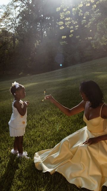 A mother in a yellow dress shares a tender moment with her toddler daughter in a sunlit green park.