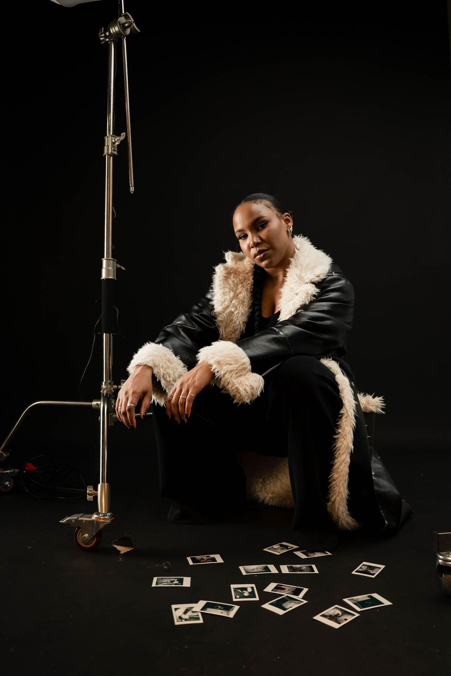Woman in black leather shearling coat sits on box surrounded by Polaroids near studio light on black background.