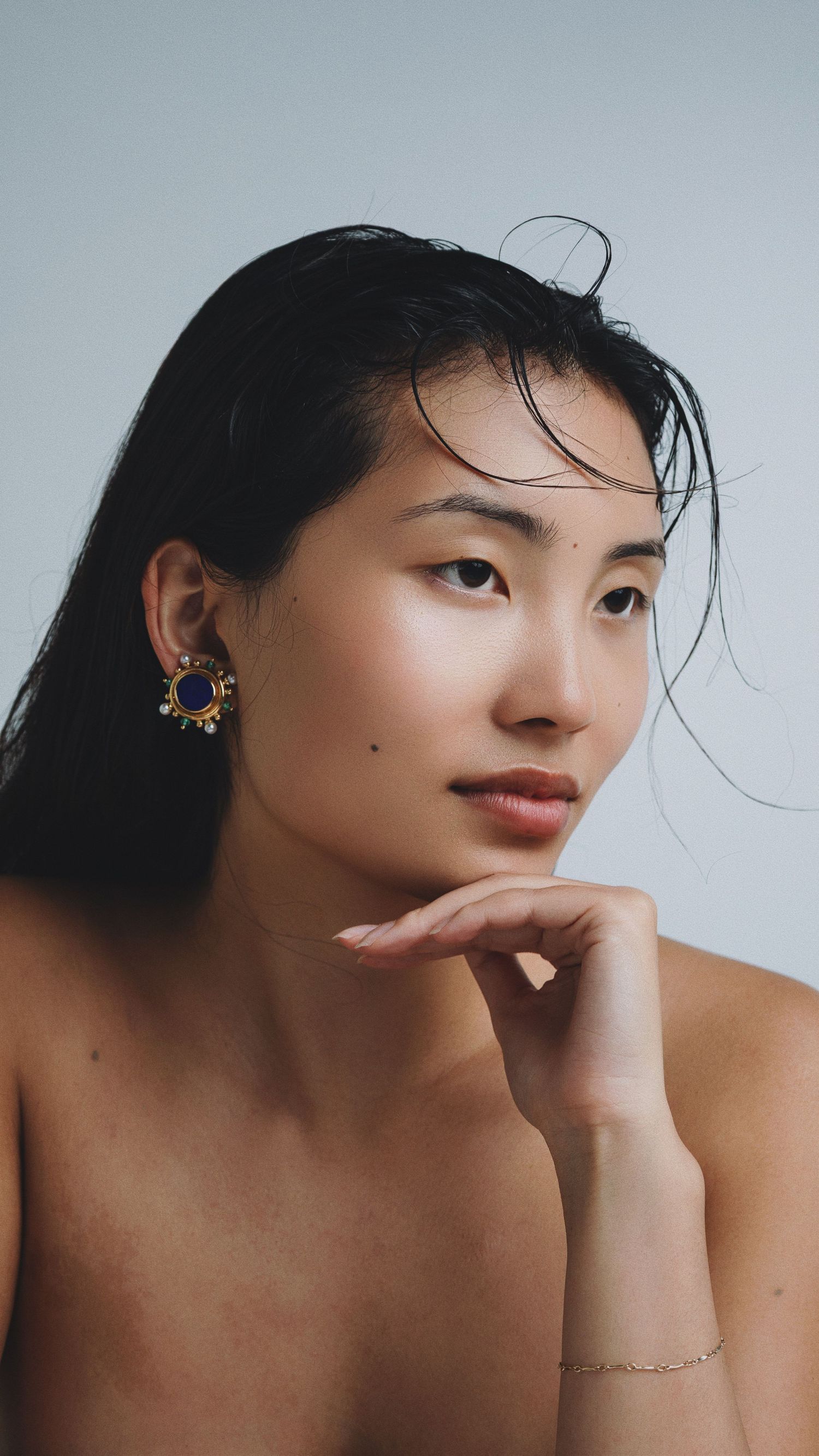 Young Asian woman with dark hair poses thoughtfully, wearing ornate gold and blue earrings and a delicate bracelet.
