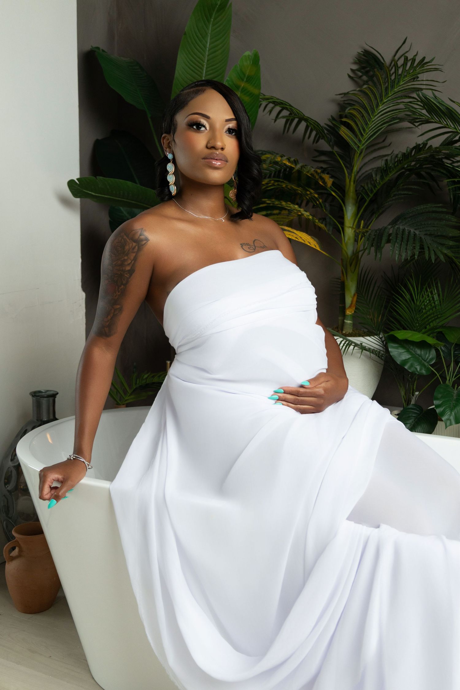 A pregnant woman in a white strapless gown sits elegantly in a bathtub surrounded by tropical plants.