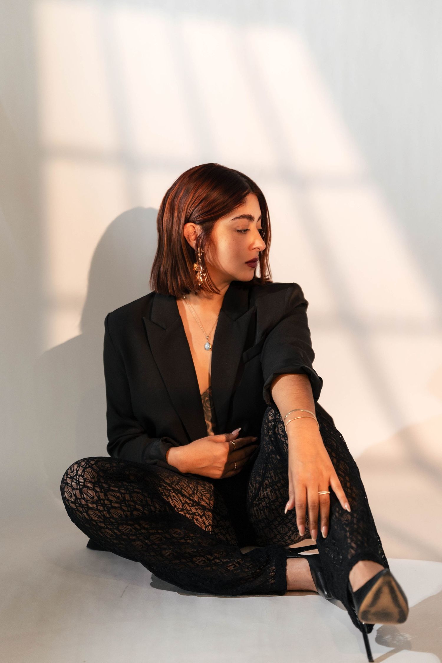 Woman in black blazer and lace pants sitting cross-legged against white wall in dramatic sunlight.
