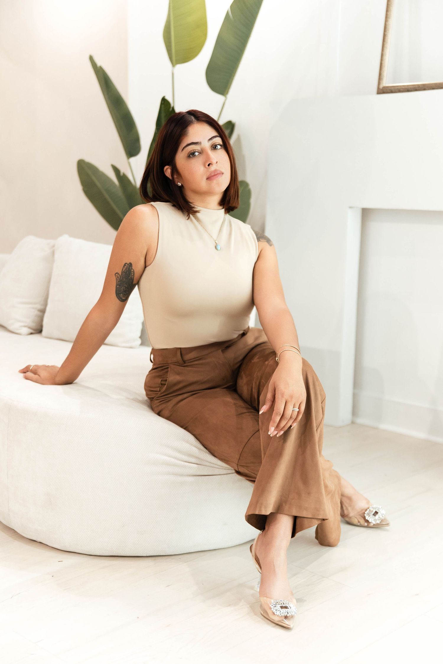 Woman in beige top and brown wide-leg pants sits on white curved sofa with plant in bright modern room.