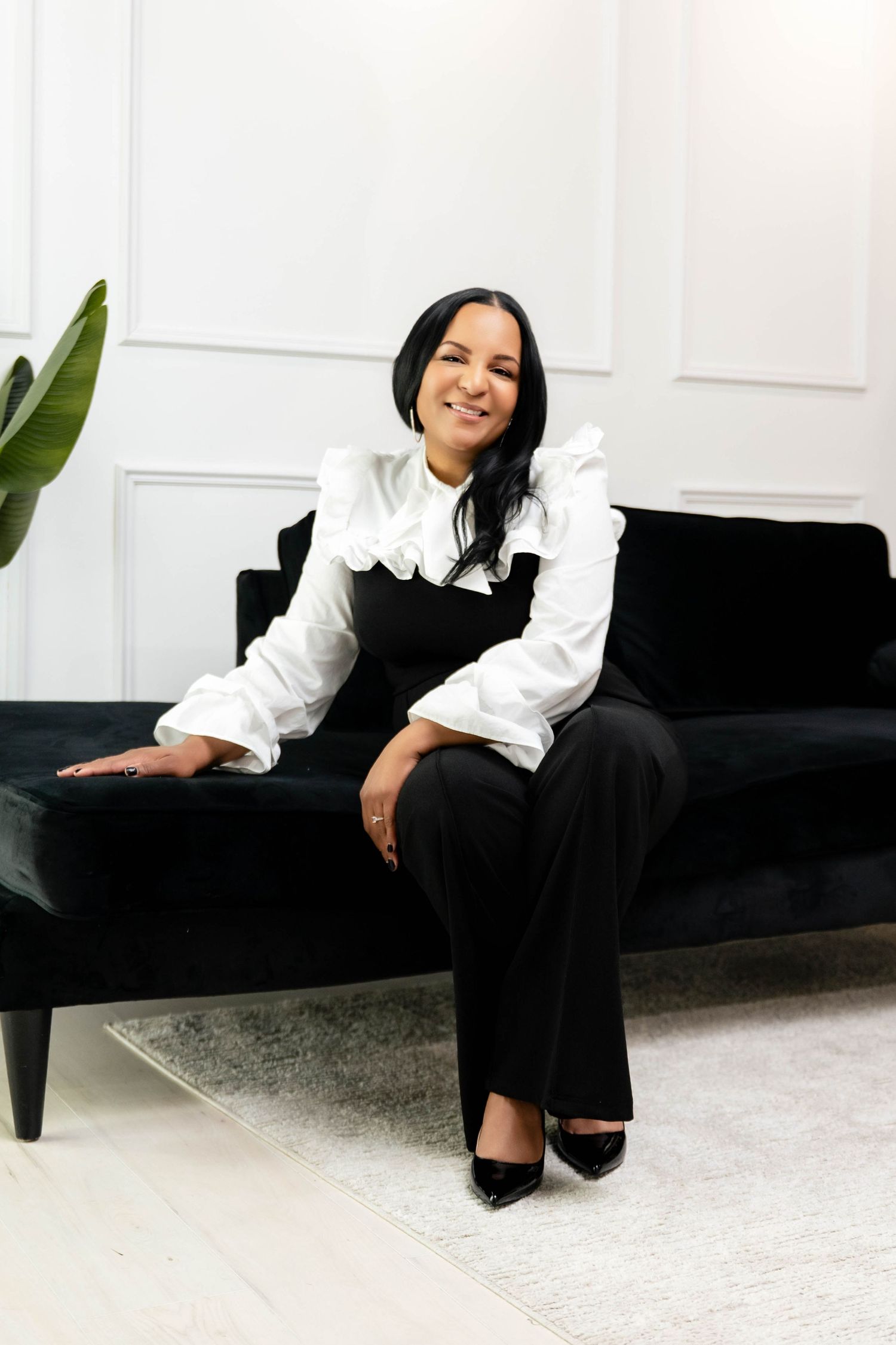 A smiling woman in a white ruffled blouse and black outfit sits elegantly on a dark velvet sofa in a bright room.