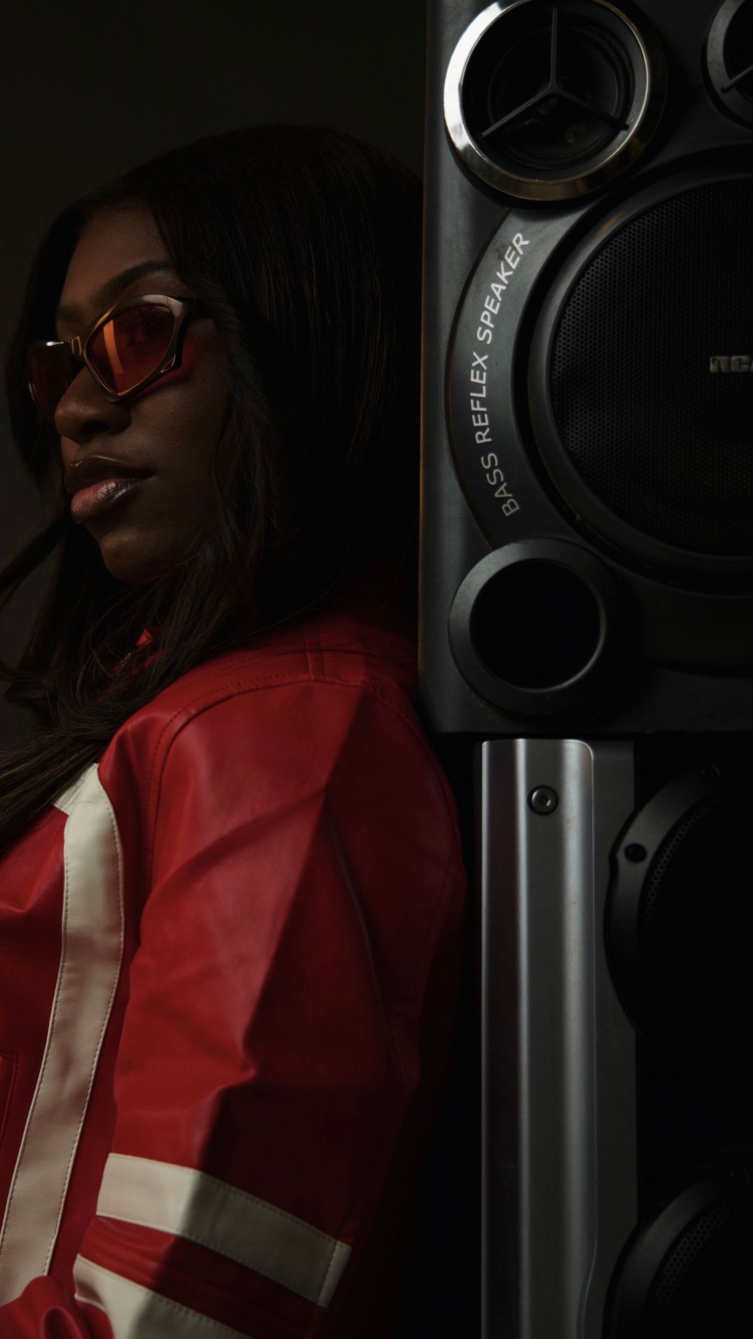 A stylish woman in a red jacket and cat-eye sunglasses poses beside a large bass reflex speaker in a dark setting.