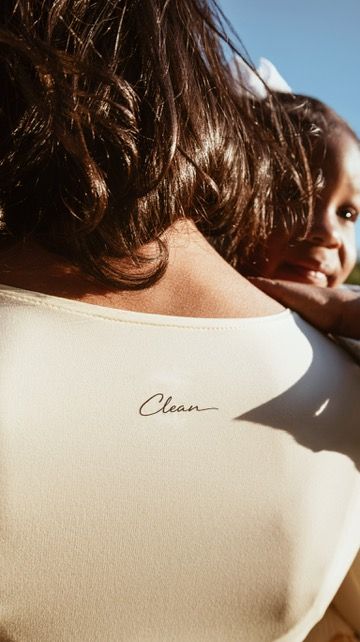 Woman in cream Clean-branded top holds smiling baby outdoors in warm sunlight.