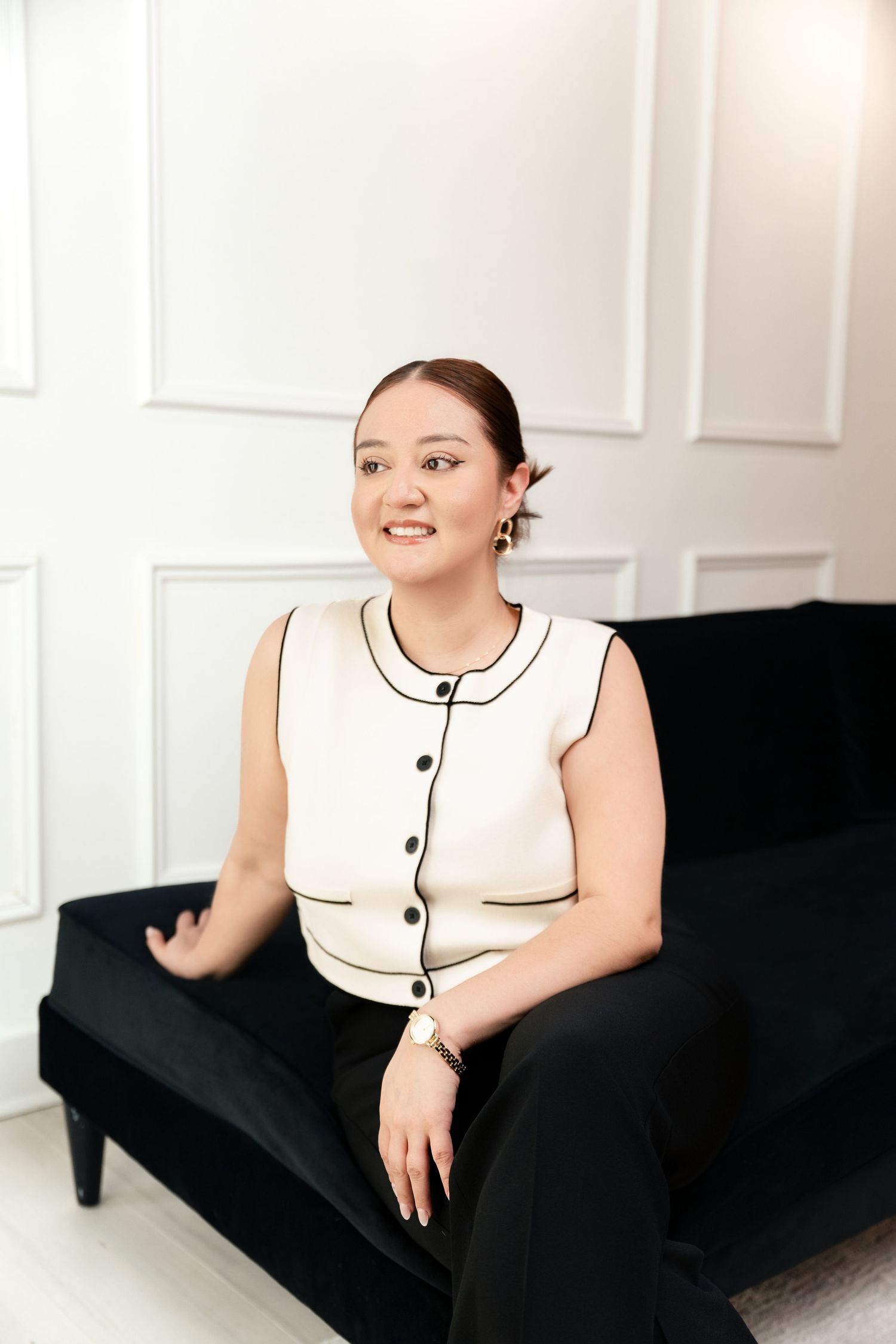 Young woman in white sleeveless top with black trim sits smiling on a dark velvet sofa against white wall.