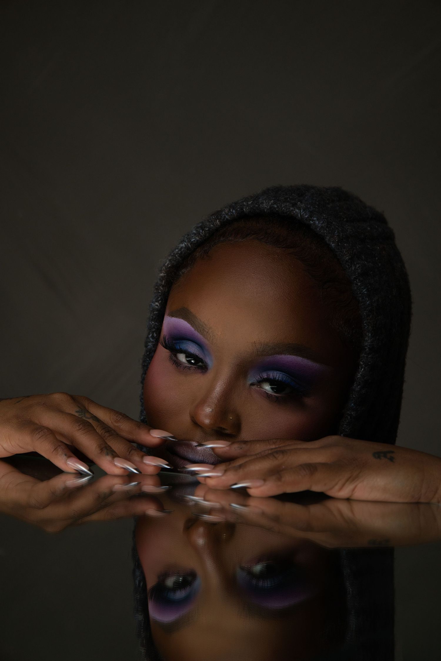 A Black woman with bold purple eye makeup and long nails rests her chin on hands, reflected on a mirror surface.